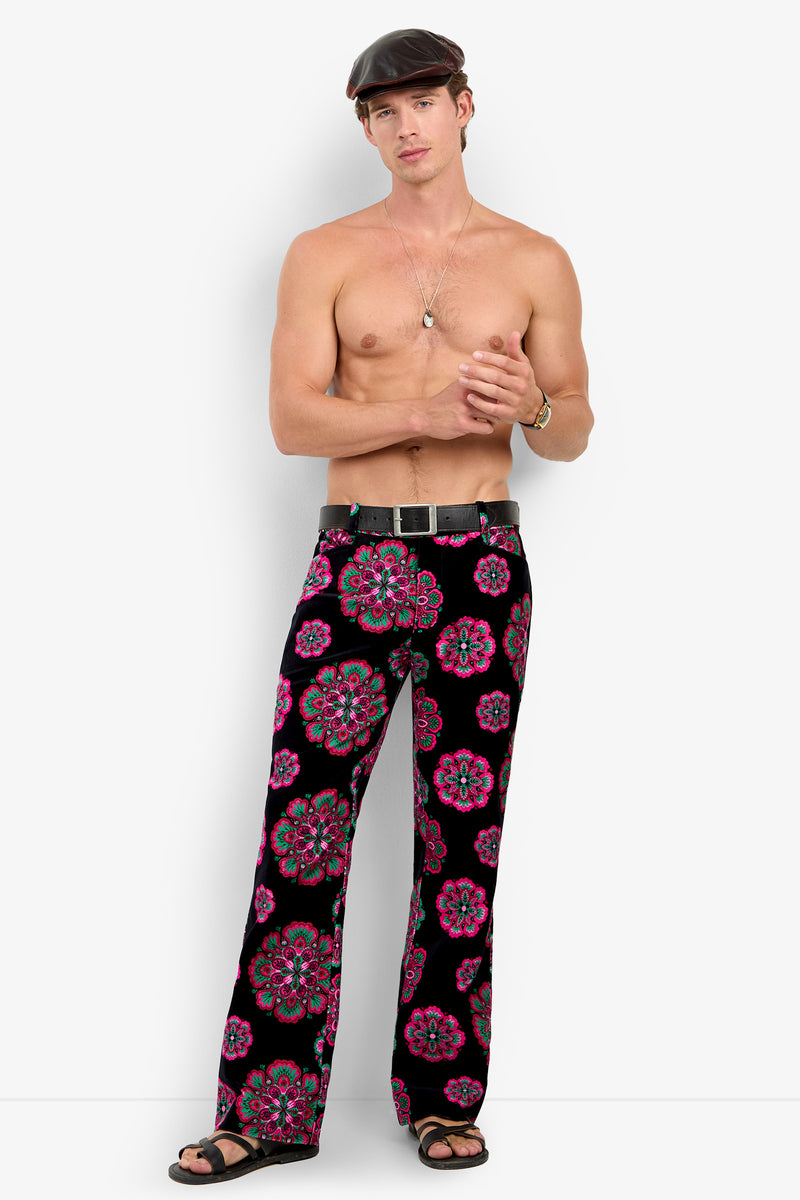 A shirtless man wearing a black flat cap, necklace, black belt, black sandals, and the Alejandro Bootleg Trouser—black pants featuring a colorful pink and green floral pattern—stands against a plain white background.