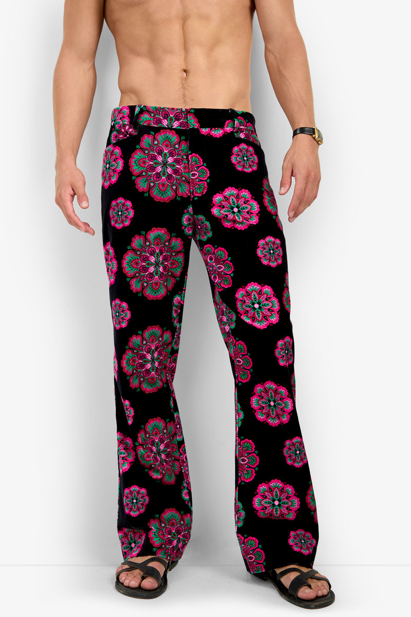 A shirtless man wears the Alejandro Bootleg Trouser—black pants with a pink and green floral pattern—along with black sandals and a wristwatch, standing against a plain white background.