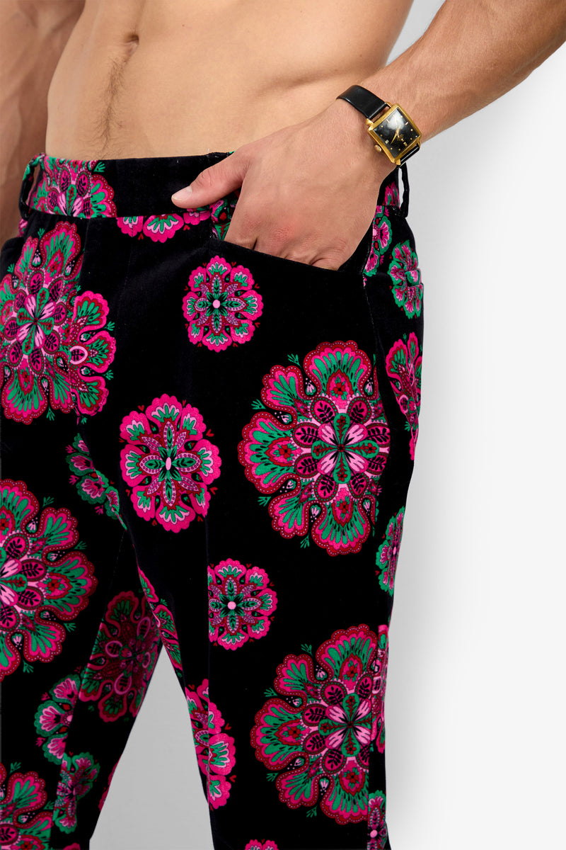 A shirtless person wearing the Alejandro Bootleg Trouser—a pair of black pants featuring vibrant pink and green floral patterns—with their left hand in their pocket and a black wristwatch visible.