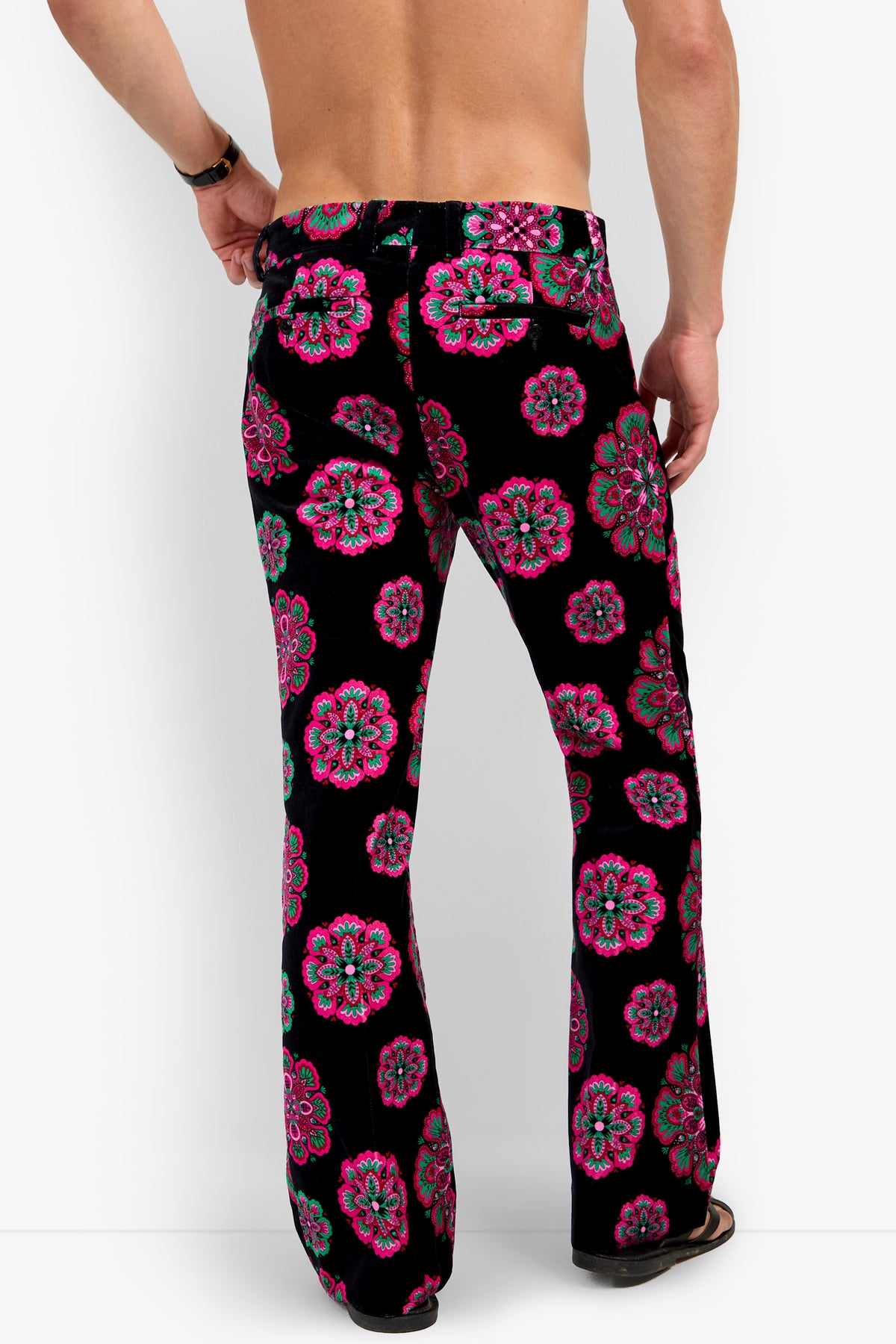 Wearing the Alejandro Bootleg Trouser, which features vibrant pink, teal, and red floral patterns on a black background, a person stands barefoot with their back and arms exposed.
