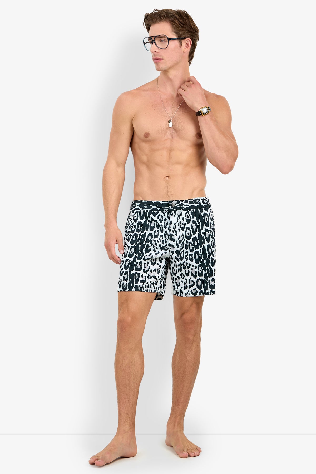 A shirtless man wearing glasses, a wristwatch, a necklace, and the HONOLULU BOARDSHORT in black-and-white leopard print stands barefoot against a plain white background.