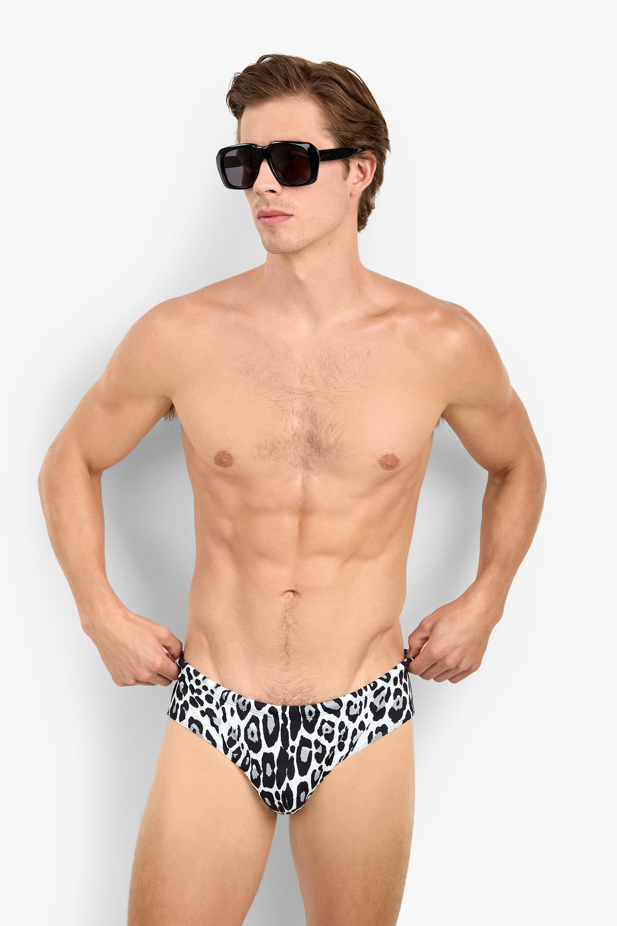 A shirtless man wearing black sunglasses and the BONDI SWIM BRIEF, featuring a black-and-white leopard print, stands against a plain white background with his hands on his hips.