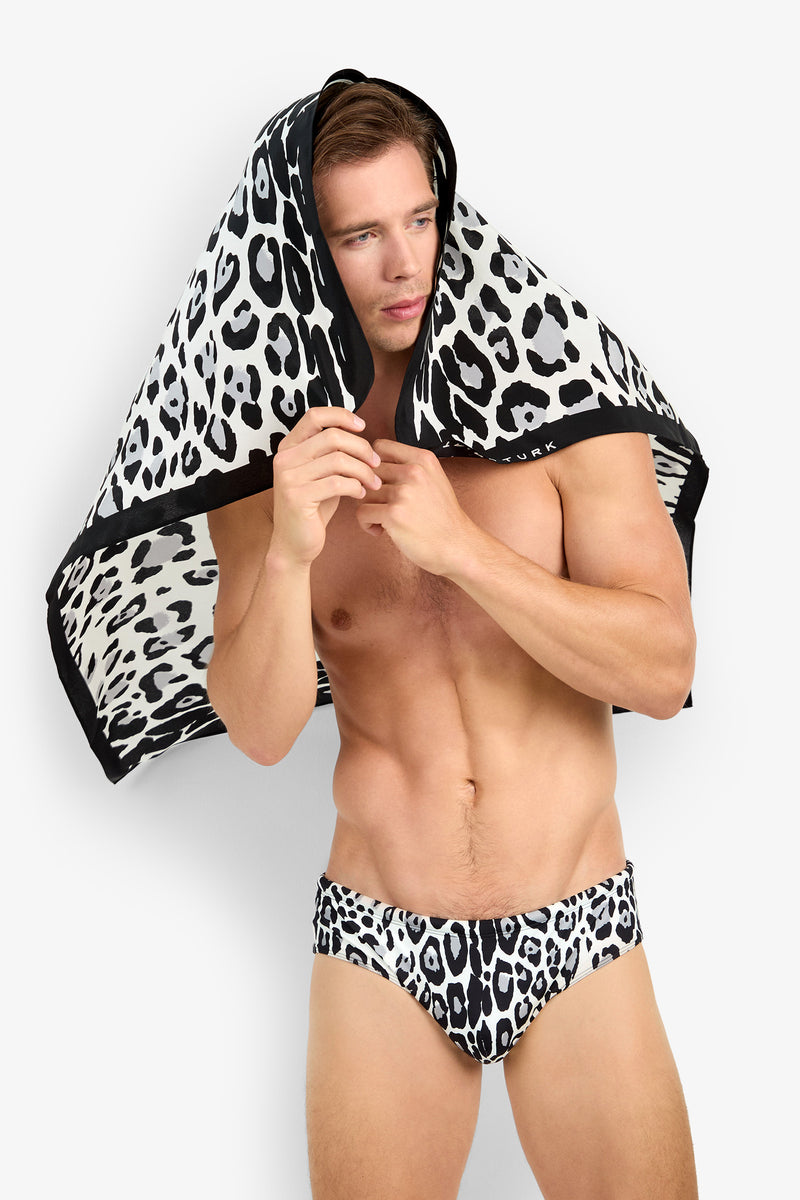 A shirtless man wears the BONDI SWIM BRIEF in leopard print and holds a matching towel over his head against a plain white background.