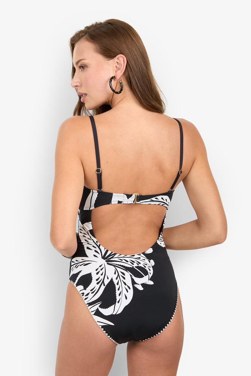 A person wearing the GOLDEN HOUR BLOOM BANDEAU ONE PIECE, a black and white floral print swimsuit with thin straps and a stylish back cutout.