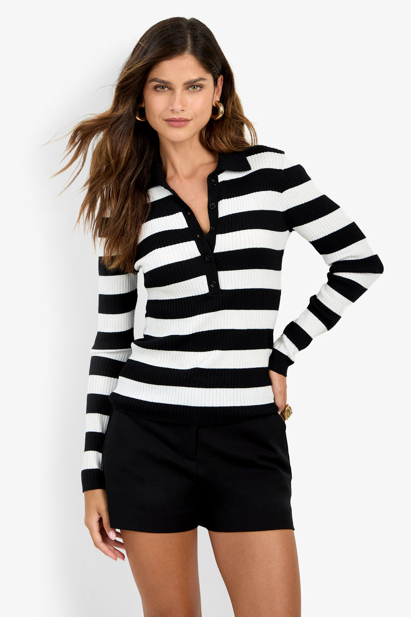 A woman wearing the Hayworth Polo, a black and white striped long-sleeve collared shirt, paired with black shorts, stands against a plain white background.