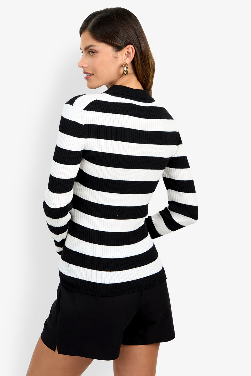 A woman wearing the Hayworth Polo, a black and white striped long-sleeve sweater, paired with black shorts stands facing away, looking over her shoulder against a plain background.