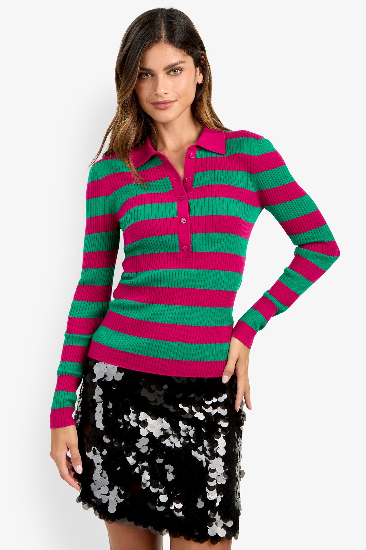 A woman is standing against a white background, smiling with one hand on her hip, wearing the Hayworth Polo—a green and pink striped long-sleeve sweater—paired with a black sequin skirt.