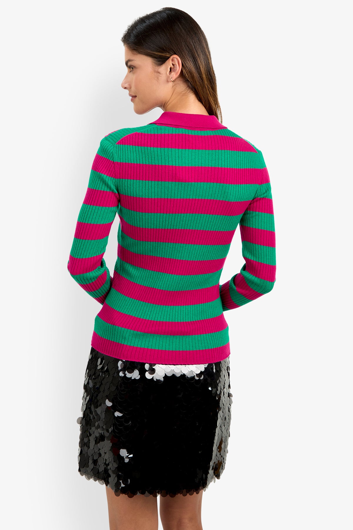 A woman is seen from behind, standing against a plain white background, wearing the Hayworth Polo—a green and pink striped sweater—paired with a black sequin-detailed skirt.
