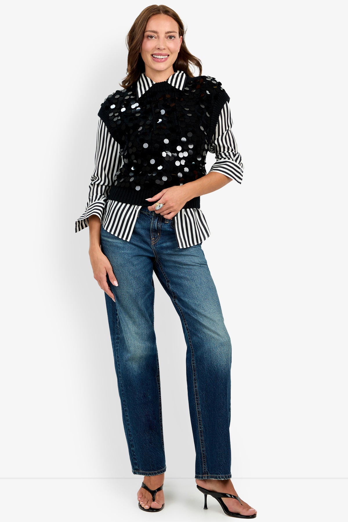 A woman in a black and white striped shirt, blue jeans, black heeled sandals, and the Dalida Vest stands smiling against a white background.