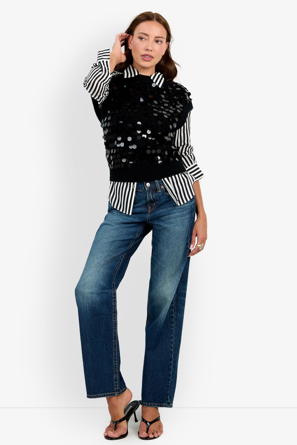 A woman stands against a plain white background with one hand touching her hair, wearing wide-leg jeans, black heeled sandals, a black-and-white striped shirt, and the Dalida Vest: a black textured vest adorned with large sequins.