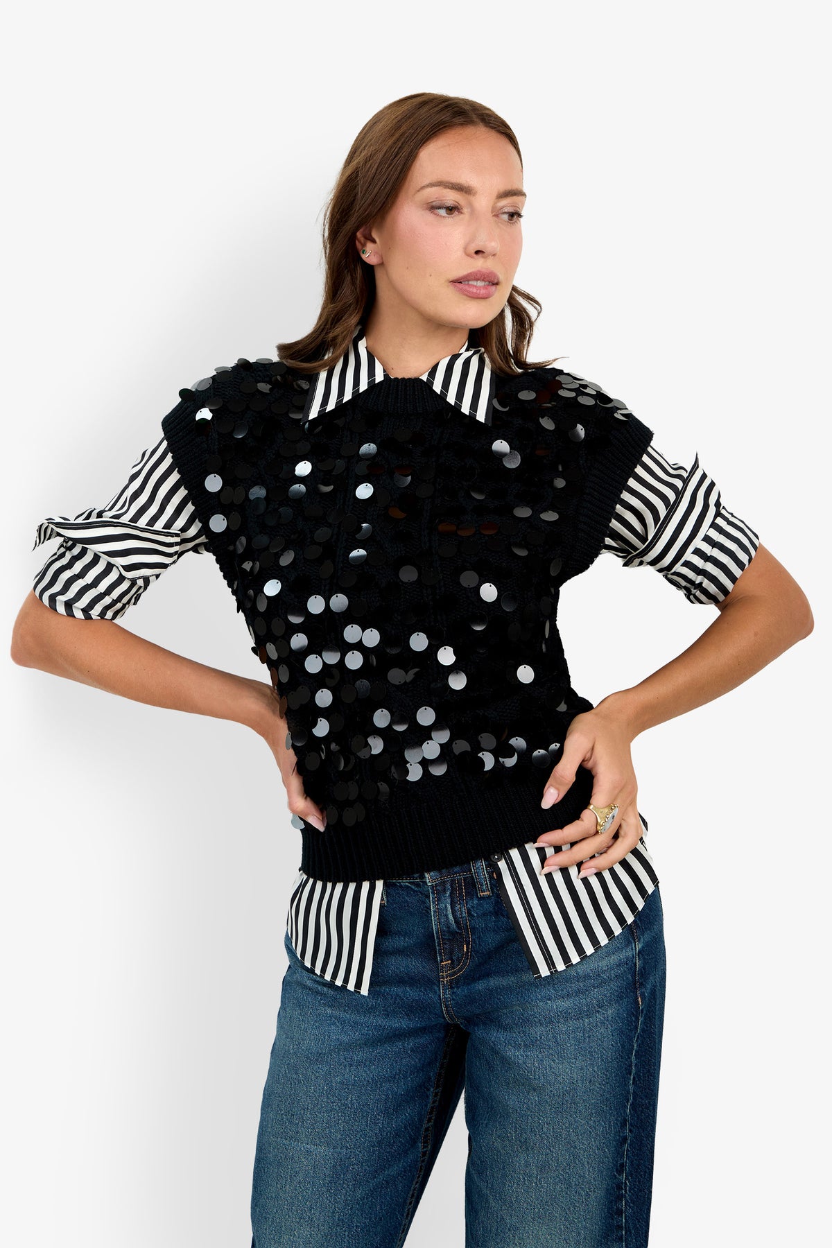A woman standing against a white background, wearing the Dalida Vest—a black, sequin-embellished piece—layered over a black-and-white striped shirt with rolled-up sleeves and paired with blue jeans, with her hands on her hips.