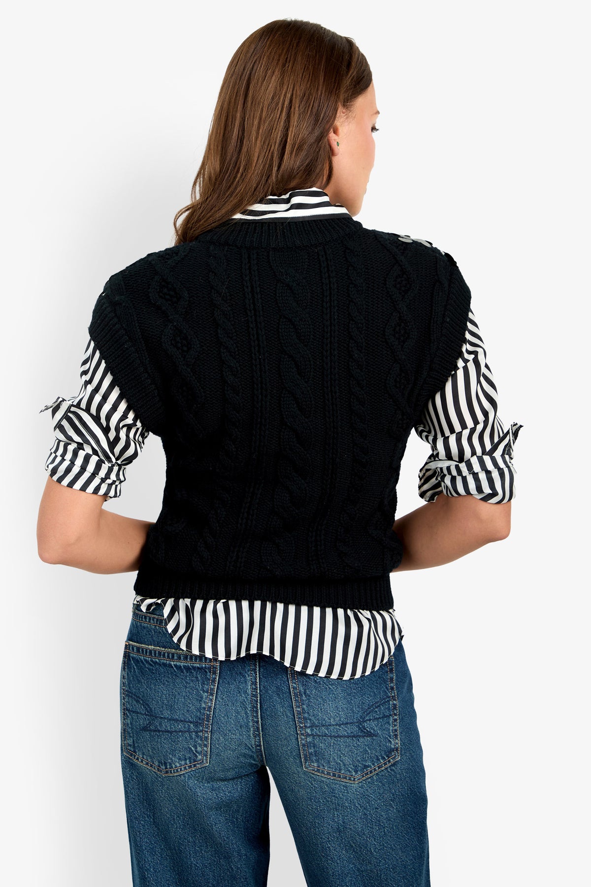 A person stands with their back to the camera, wearing the Dalida Vest—a black cable knit piece—over a black-and-white striped shirt and blue jeans.