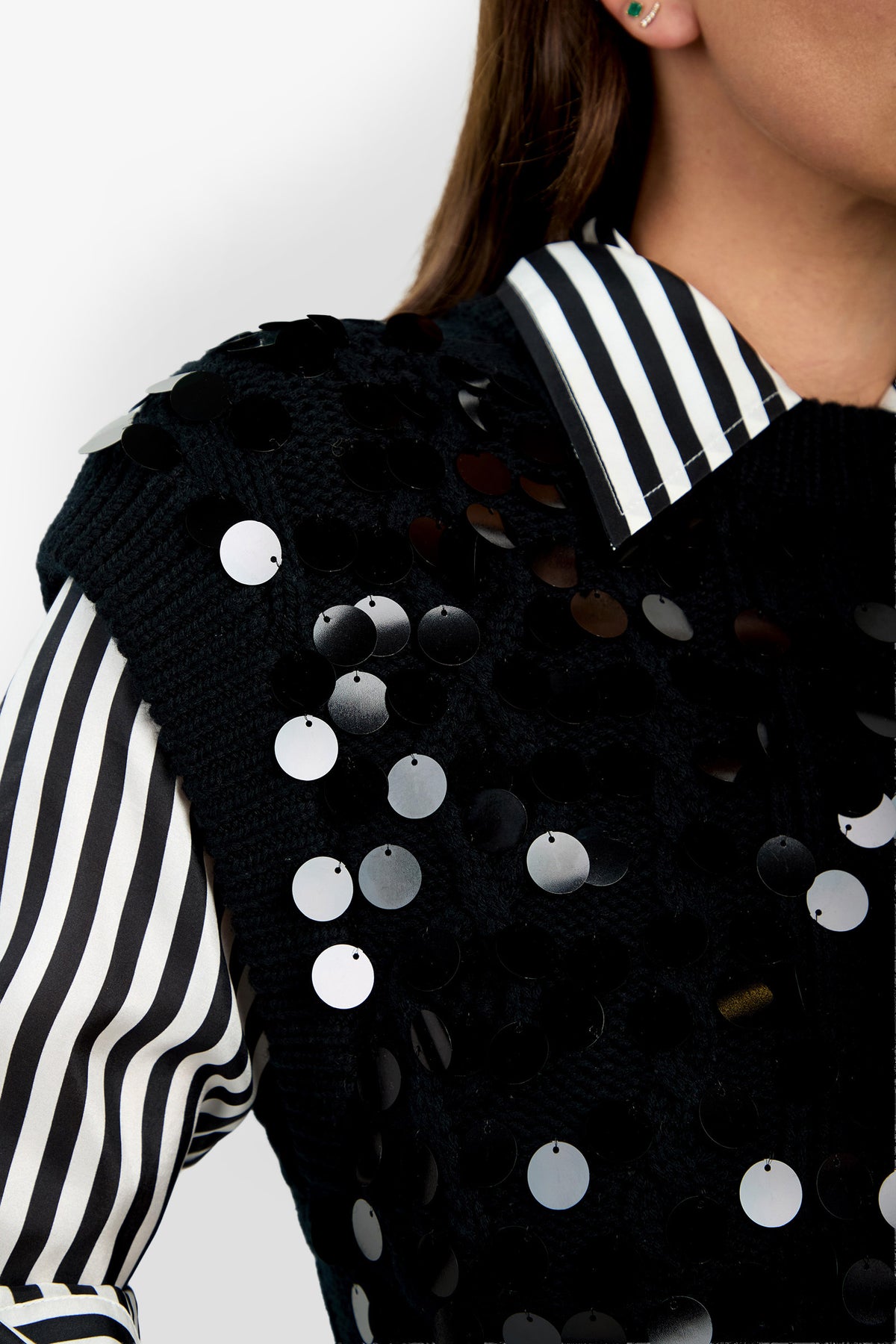 A person wearing the Dalida Vest—a black knitted vest adorned with shiny black sequins—layered over a black-and-white vertical striped collared shirt; only part of the upper body and face are visible.