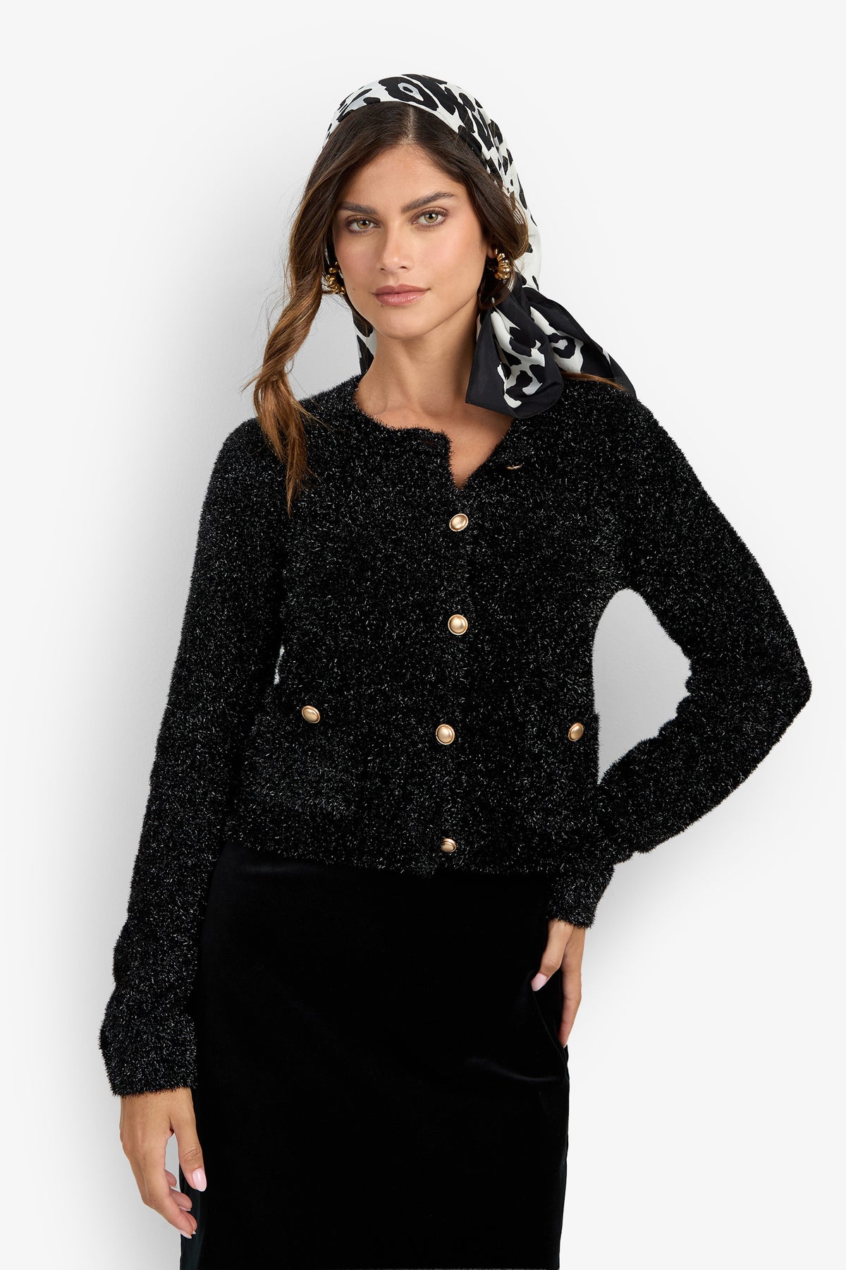 A woman wearing the Sinai Cardigan—a fuzzy black button-up sweater—paired with a black skirt and a black-and-white patterned scarf in her hair, stands against a plain light background.