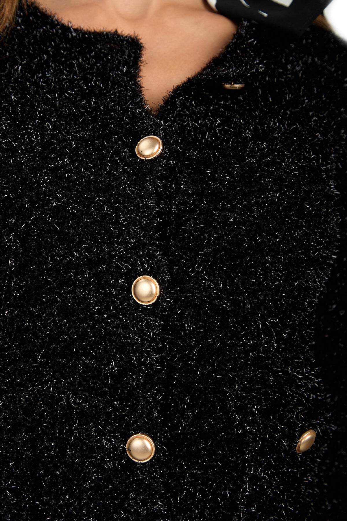 Close-up of a person wearing the Sinai Cardigan, a textured black fuzzy jacket with shiny gold buttons, and a small part of a black and white patterned scarf visible at the neckline.