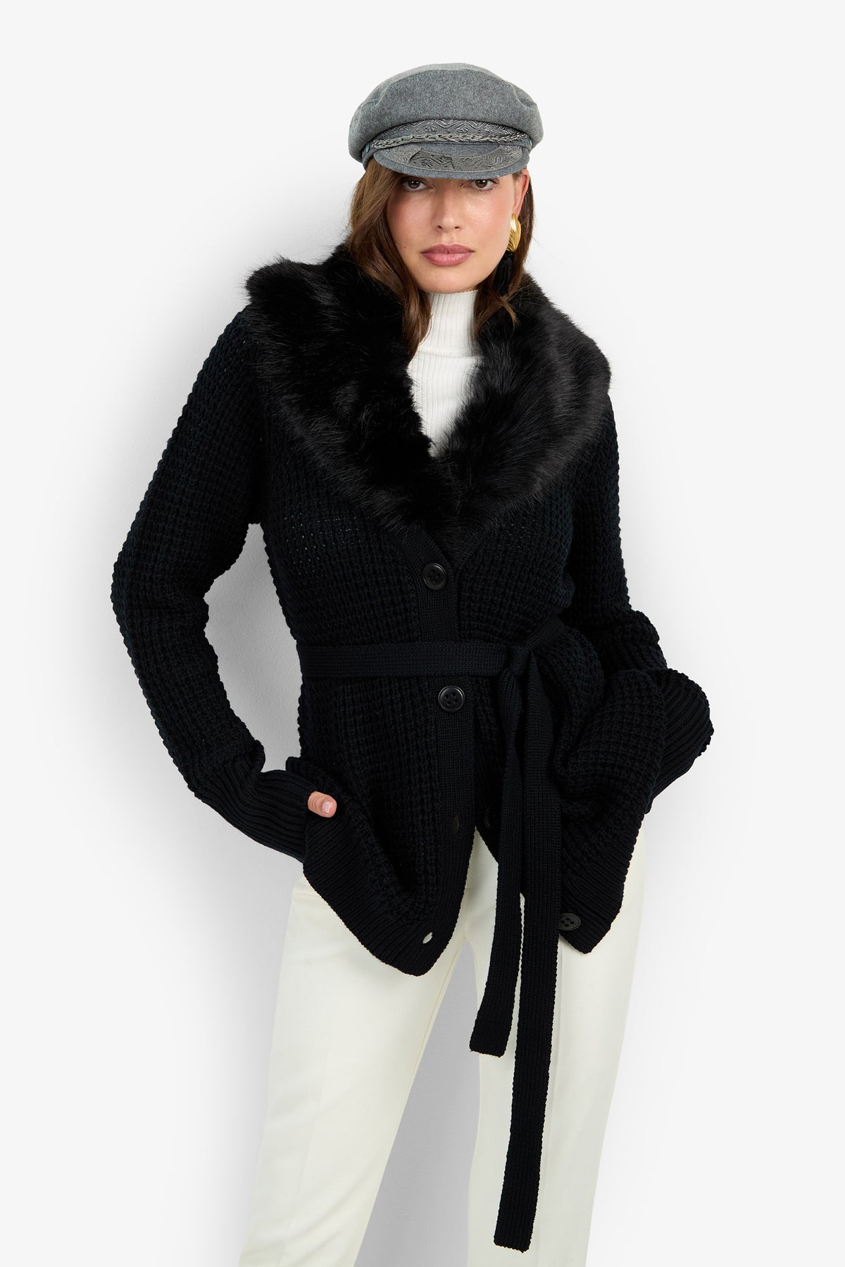 A woman wearing the Pellicia Fur Cardigan—a black belted knit cardigan with a faux fur collar—paired with a gray newsboy cap, white turtleneck, and white pants stands against a plain white background.