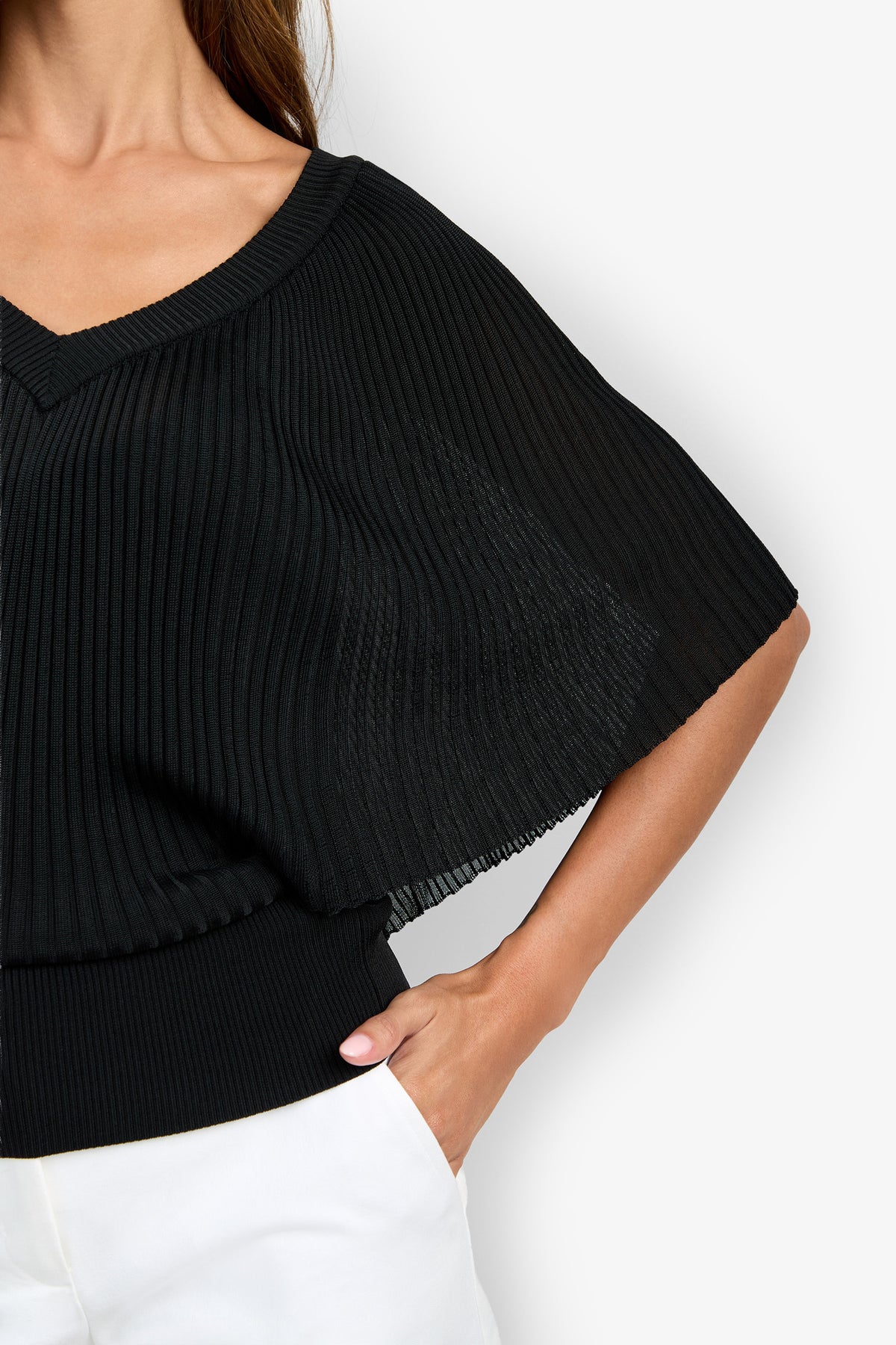 A woman wearing the Nanette Pullover—a black, pleated, short-sleeve top with a V-neckline—paired with white pants, with one hand in her pocket on a light background.