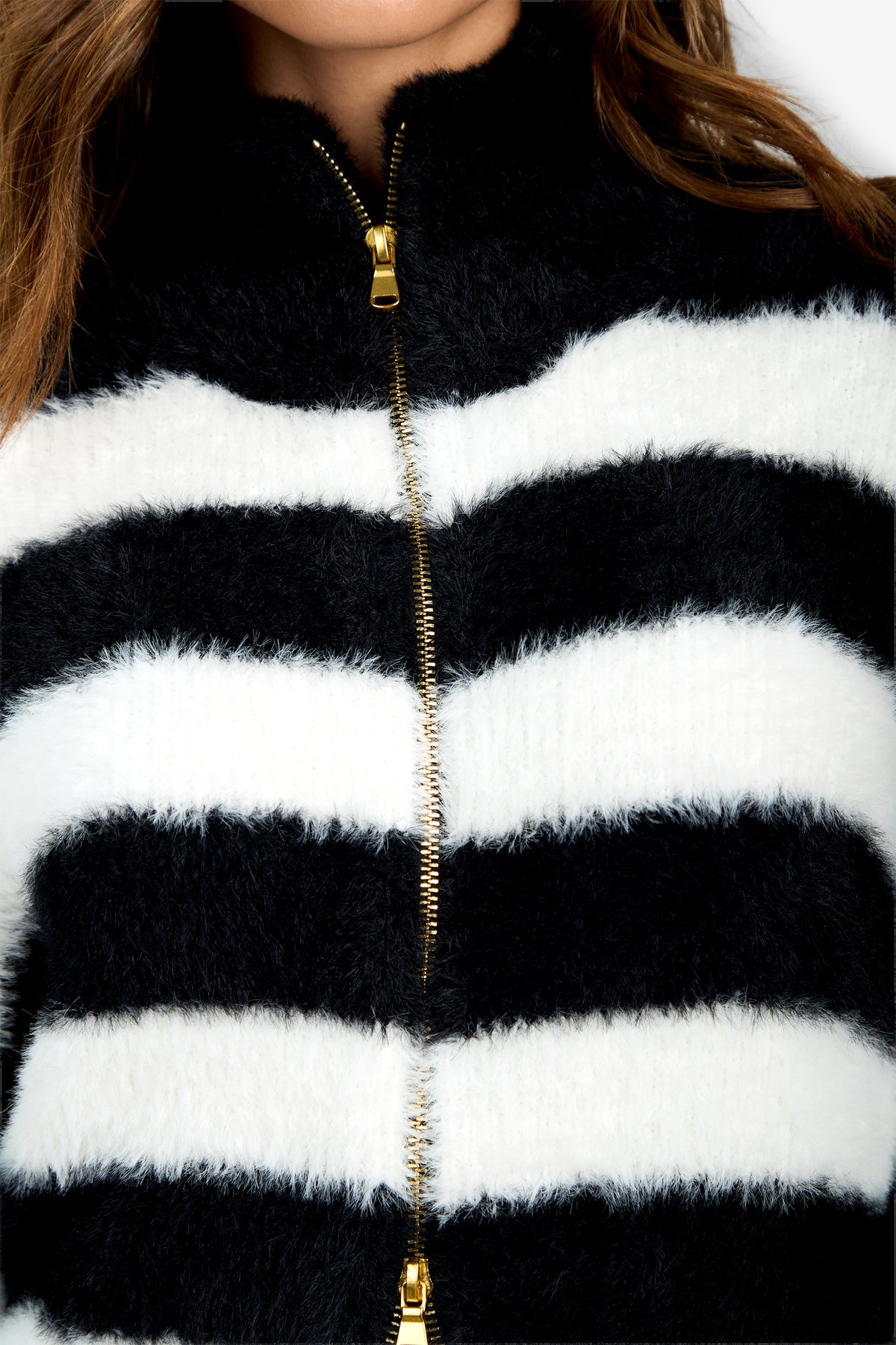 A close-up of a person wearing the Raja Sweater—a fuzzy black and white striped jacket with a gold zipper down the center. Only the lower face, hair, and upper body are visible.
