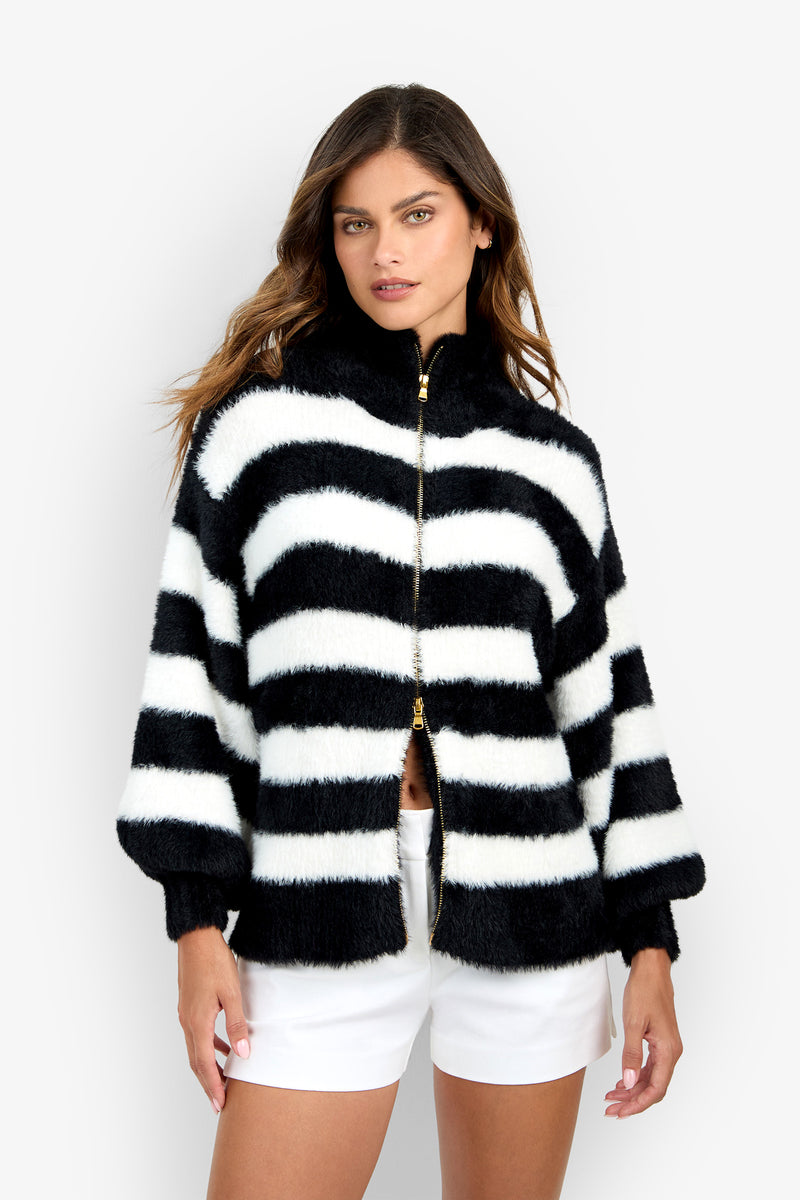 A woman wearing the Raja Sweater—a fuzzy, black and white striped zip-up jacket—paired with white shorts, stands against a plain white background.