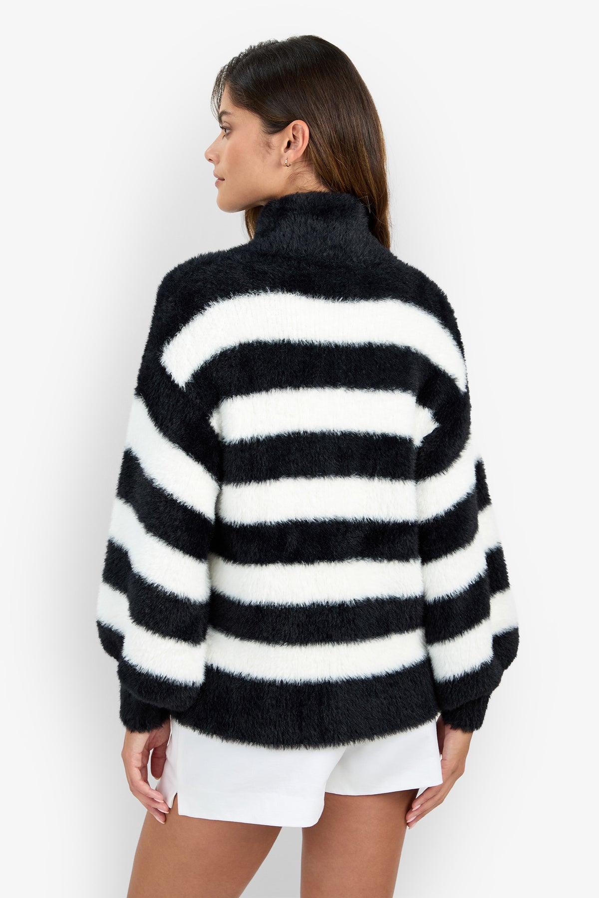 A woman facing away wears the Raja Sweater, a fuzzy black and white striped sweater with a high collar, paired with white shorts against a plain white background.