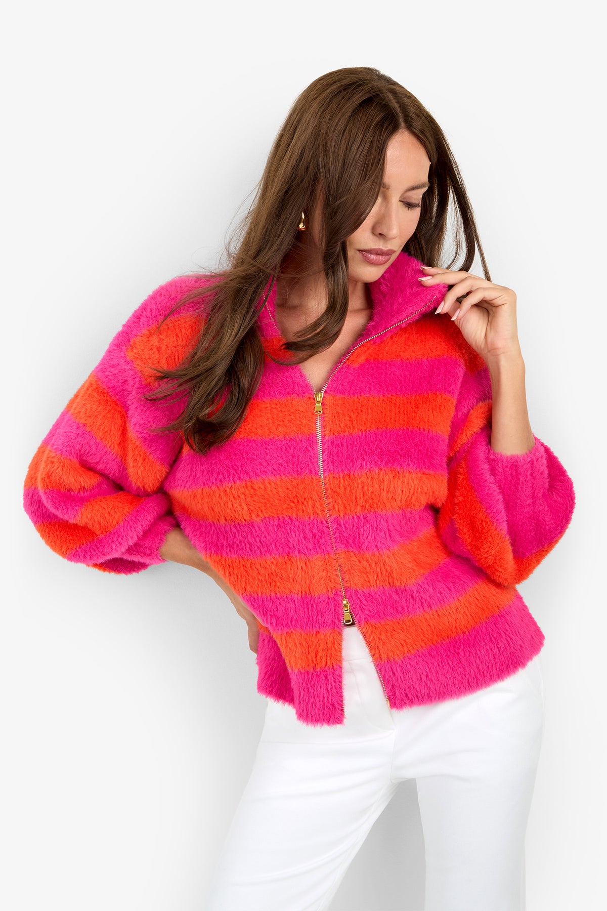 A woman wearing the Raja Sweater—a fuzzy, zip-up jacket featuring bold pink and orange horizontal stripes—paired with white pants poses against a plain background, looking down with one hand touching her collar.