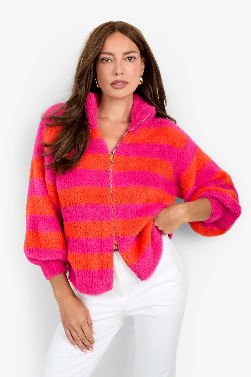 A woman wears the Raja Sweater—a fuzzy, zip-up jacket featuring bold pink and orange horizontal stripes—paired with white pants, standing against a plain white background.