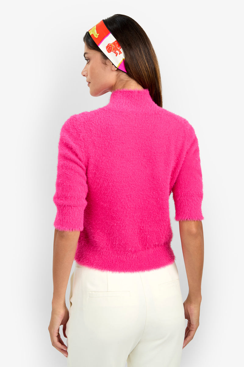 A woman is facing away, wearing the Mali Pullover—a fuzzy bright pink sweater with half sleeves—paired with white pants and a colorful red, yellow, and white headband. The background is plain white.
