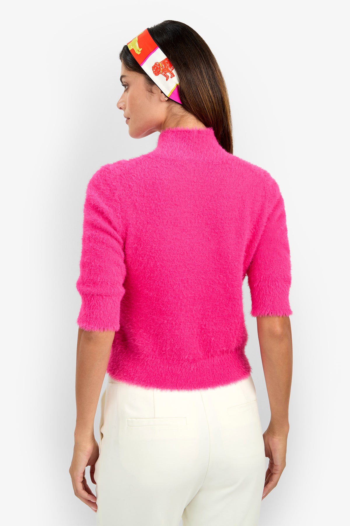 A woman is facing away, wearing the Mali Pullover—a fuzzy bright pink sweater with half sleeves—paired with white pants and a colorful red, yellow, and white headband. The background is plain white.