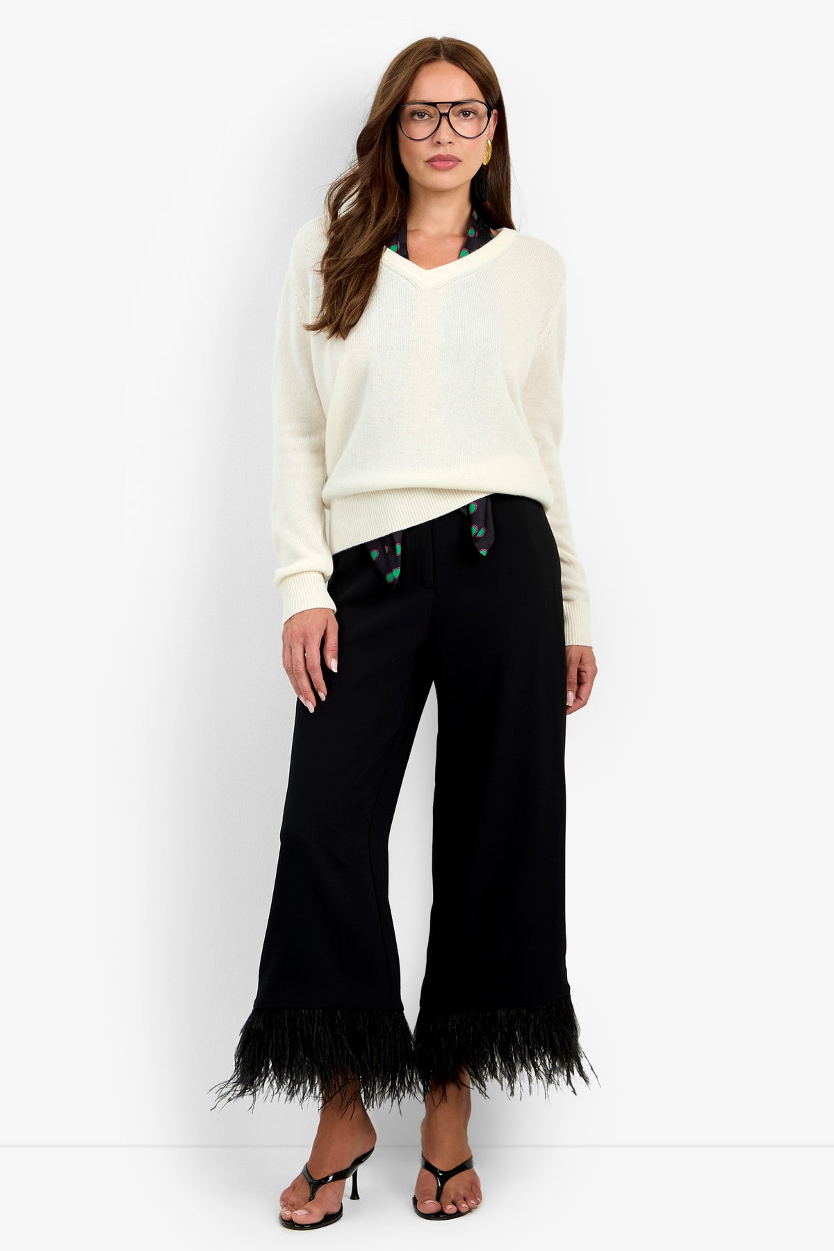 A person wears a luxury cashmere cream V-neck EVANGELINE SWEATER over a dark patterned scarf with black wide-leg trousers that have a feathered hem and black heeled sandals.