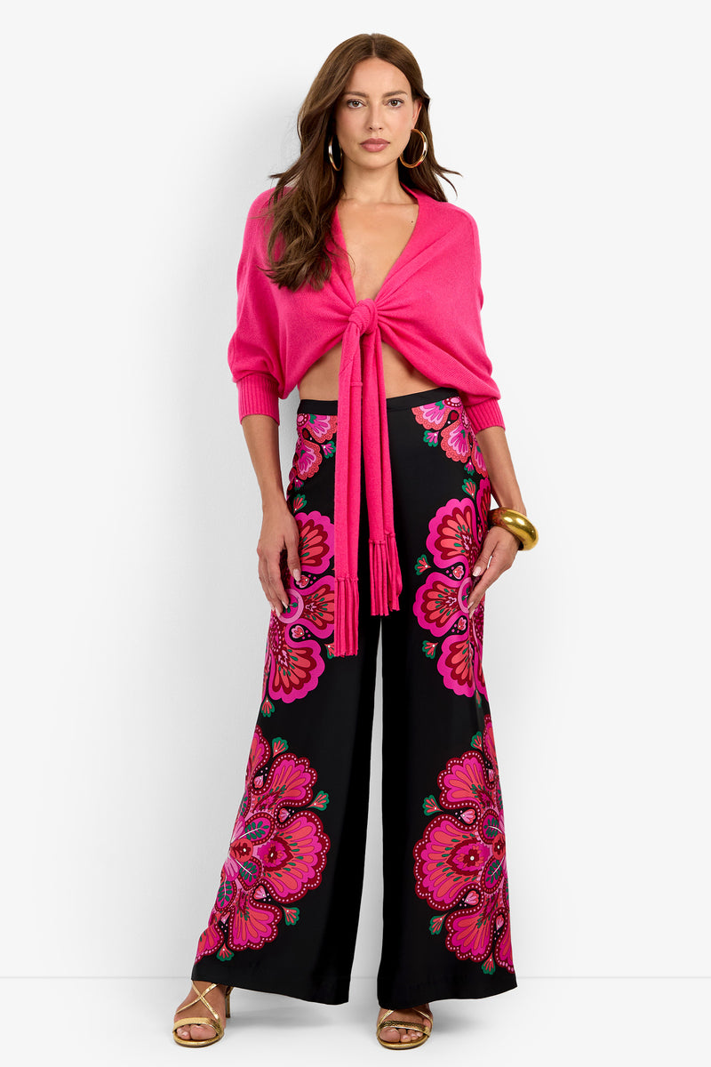 A person wearing a bright pink luxury cashmere FLORENTINA WRAP tie-front cardigan and black wide-leg trousers with a pink and magenta floral print.