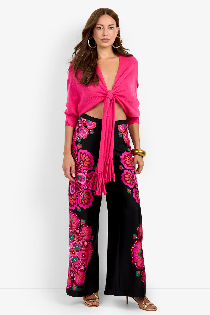 A woman wears a FLORENTINA WRAP in bright pink 100% cashmere with a tie-front, paired with wide-leg trousers featuring a large-scale pink and black floral pattern.