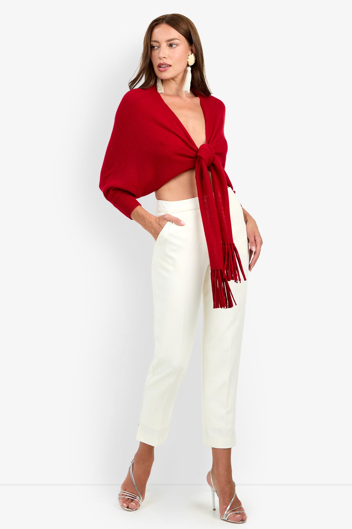 A woman wears a red cashmere FLORENTINA WRAP cropped sweater with front tassels, paired with slim-fit cream trousers and silver strappy heels.