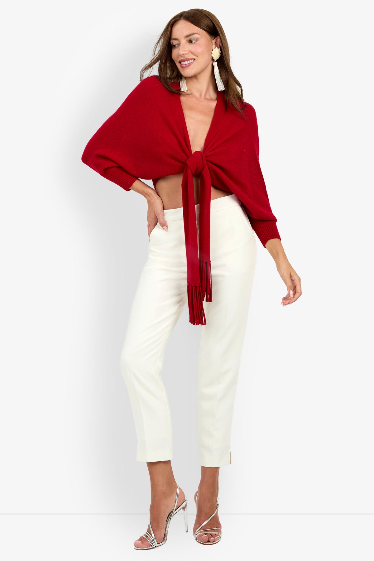 A woman wears a deep red luxury cashmere FLORENTINA WRAP top with long sleeves tied at the center, paired with cream-colored slim-fit trousers and silver high-heeled sandals.