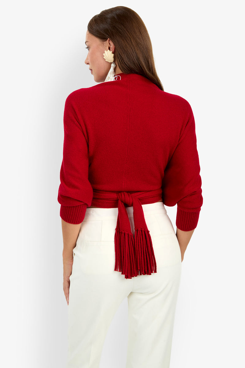 A woman seen from the back wearing a red long-sleeved FLORENTINA WRAP cashmere sweater with a tasseled hem tied at the waist over white trousers.