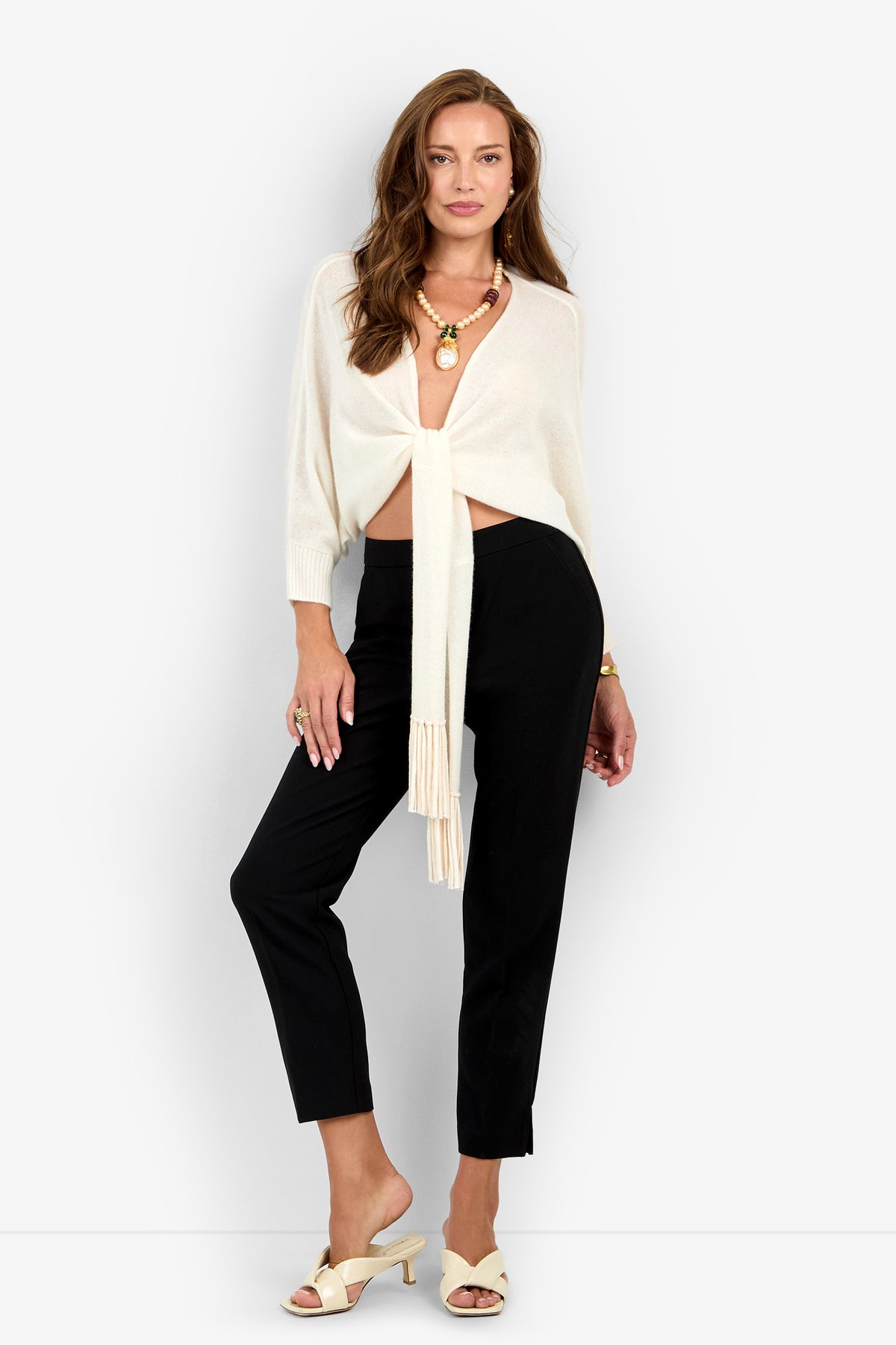 A woman wearing a cream-colored Florentina Wrap long-sleeved top that ties at the waist, black trousers, and cream-colored mules.