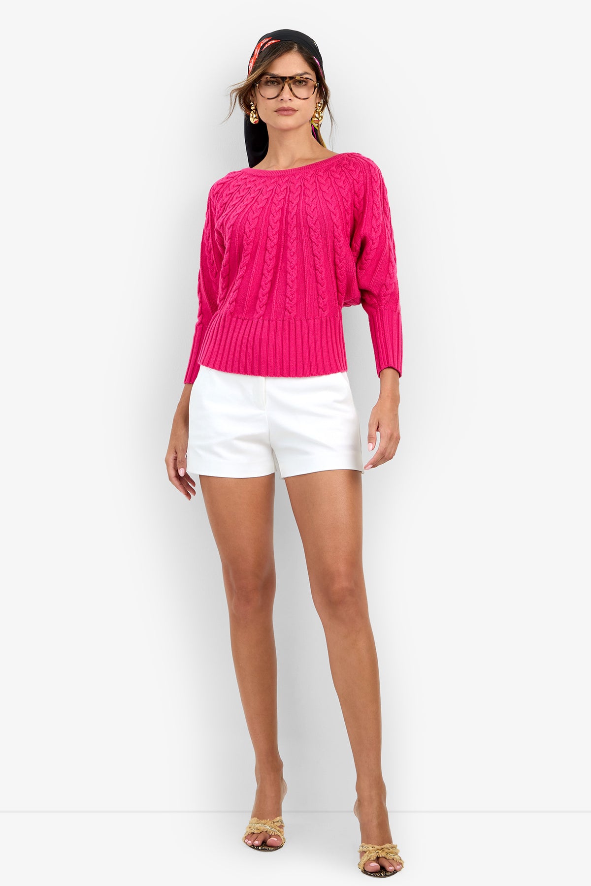 A woman wears a bright pink Tyson Knit Sweater with white shorts and heeled sandals.