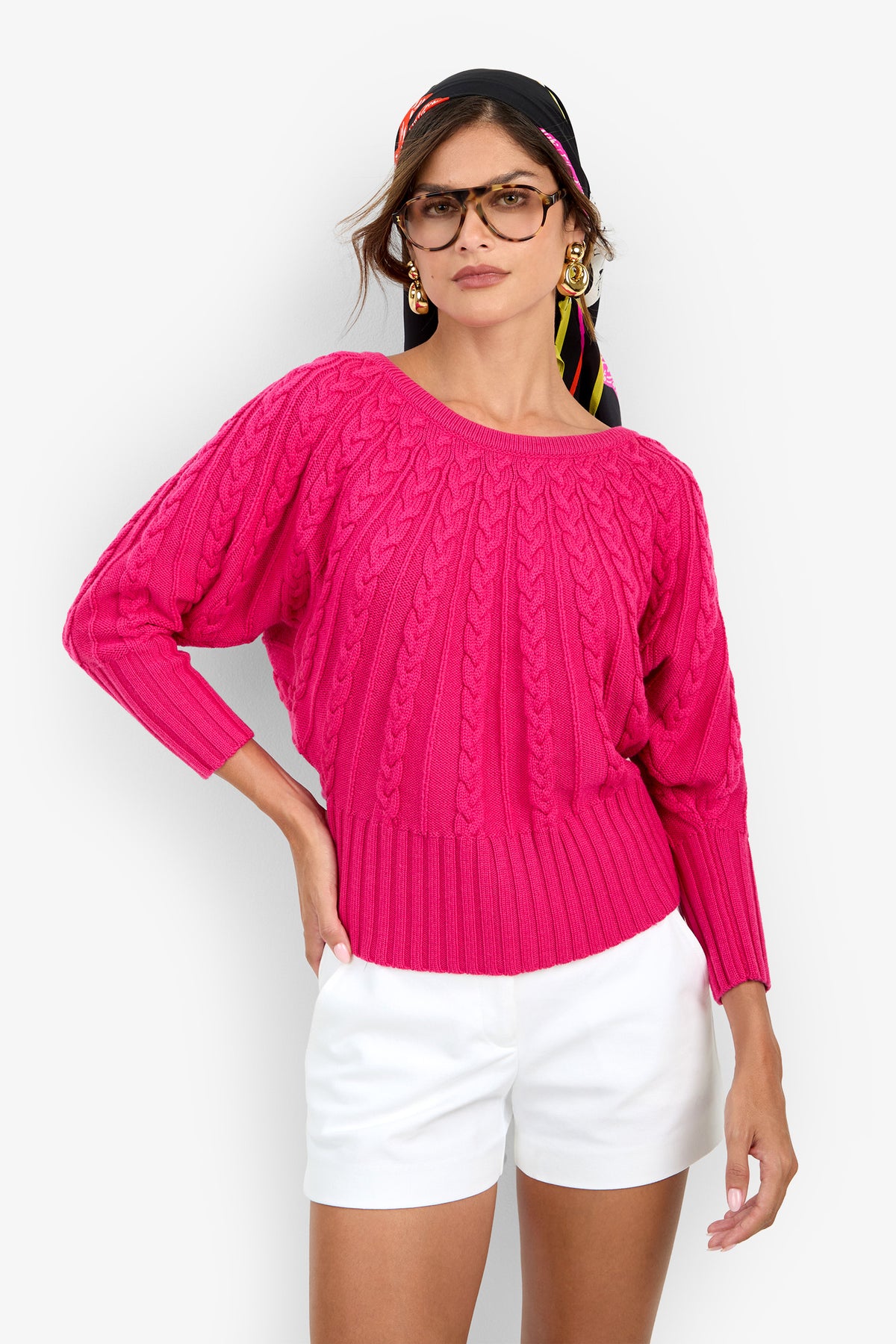 A person wears a hot pink Tyson Knit Sweater featuring a cable-knit pattern with a wide ribbed hem and cuffs, paired with white shorts.