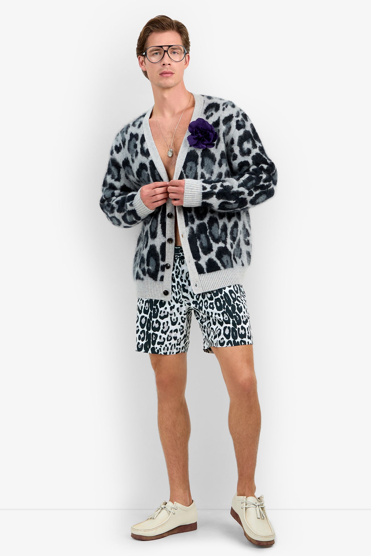 A person wearing glasses, the LEO FUZZY CARDIGAN in snow leopard, matching shorts, white shoes, a necklace, and a purple flower pin stands facing forward against a plain white background.