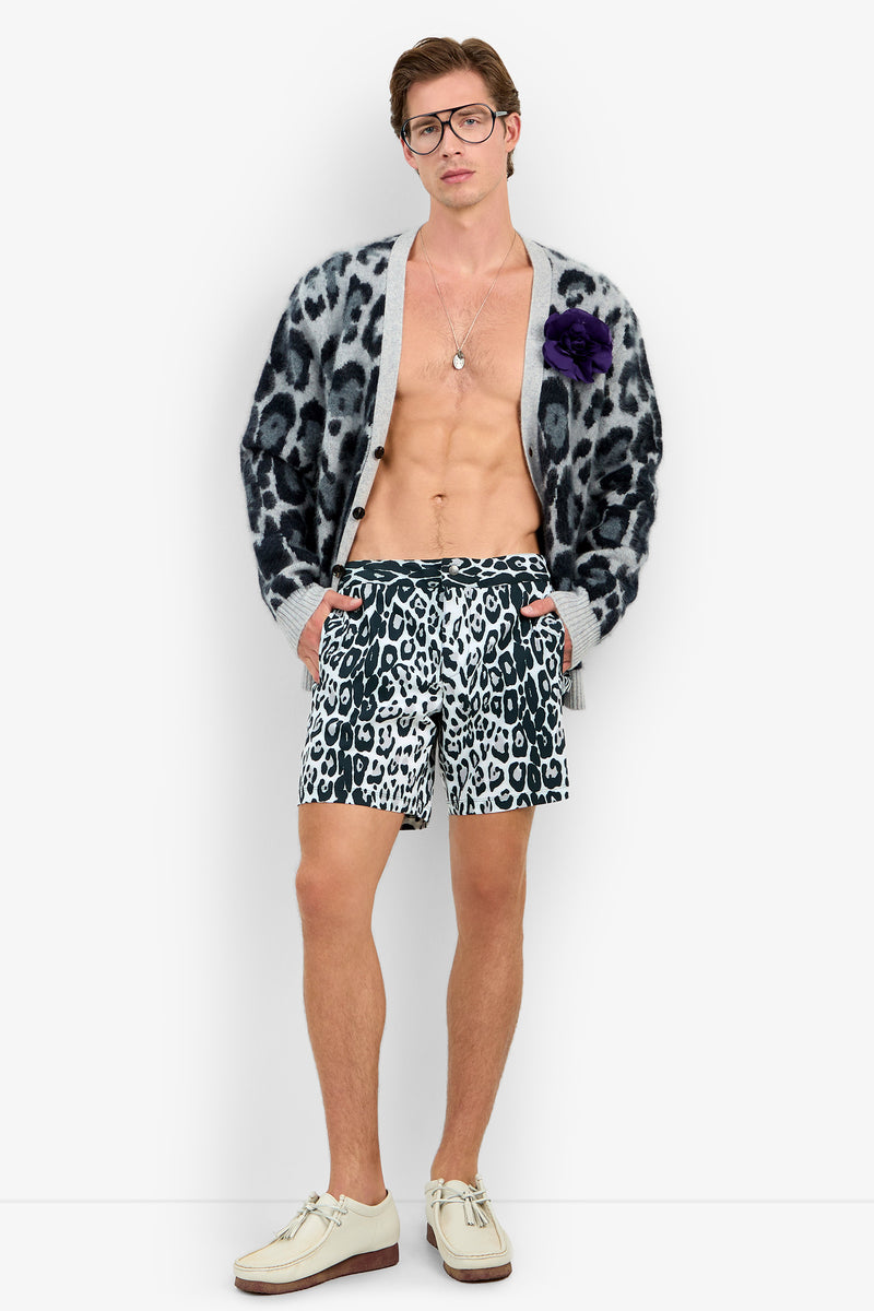 A man wearing glasses and the LEO FUZZY CARDIGAN — an open leopard-print cardigan featuring a purple flower on the chest — with matching leopard-print shorts stands barefoot against a plain white background.