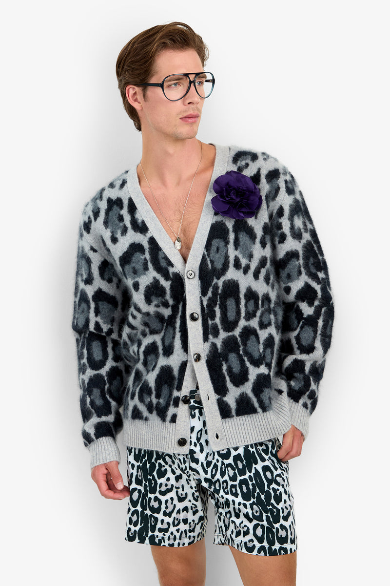 A man wearing large glasses, the LEO FUZZY CARDIGAN with a black and gray leopard print, matching animal print shorts, and a purple flower pinned to his cardigan stands against a white background.