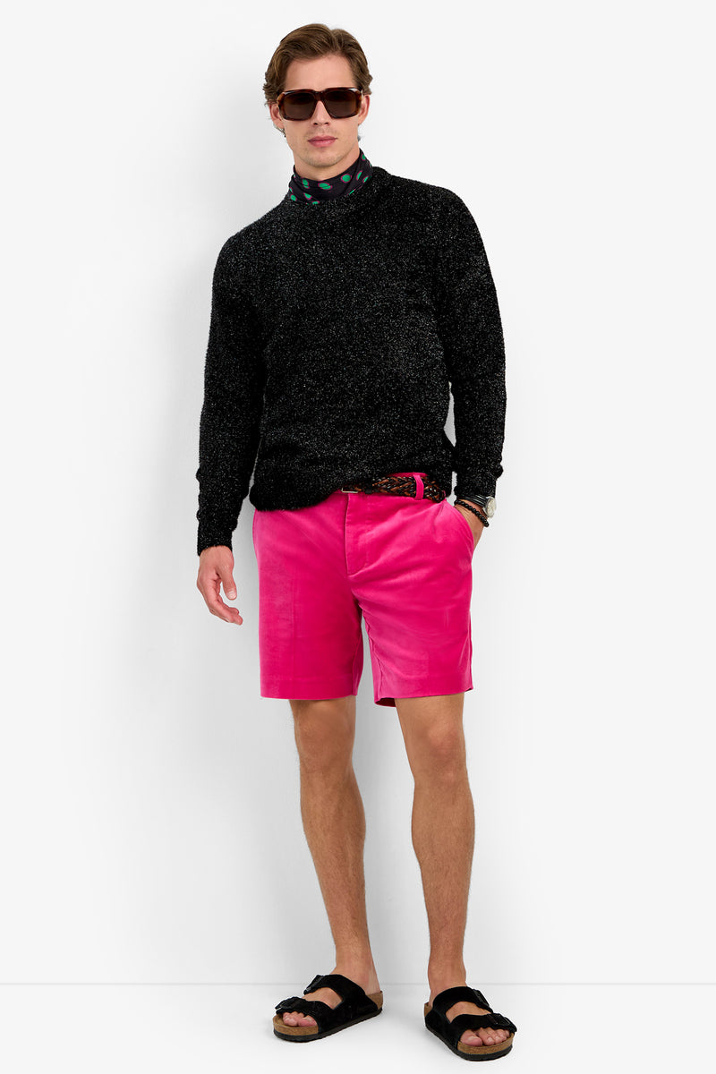 A man wearing the ASTRON SPARKLE CREWNECK in black, paired with pink shorts, black sandals, sunglasses, and a patterned shirt underneath stands against a white background with one hand in his pocket.