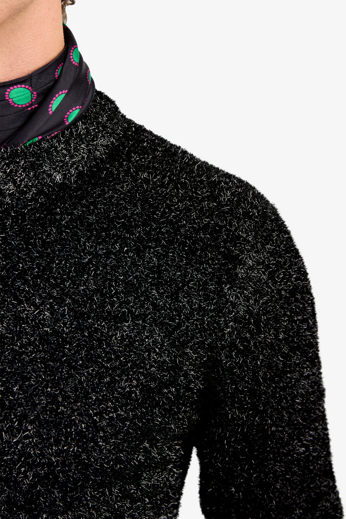 Close-up of a person wearing the ASTRON SPARKLE CREWNECK—a fuzzy black sweater—paired with a black scarf featuring green and pink circular patterns, against a plain white background.