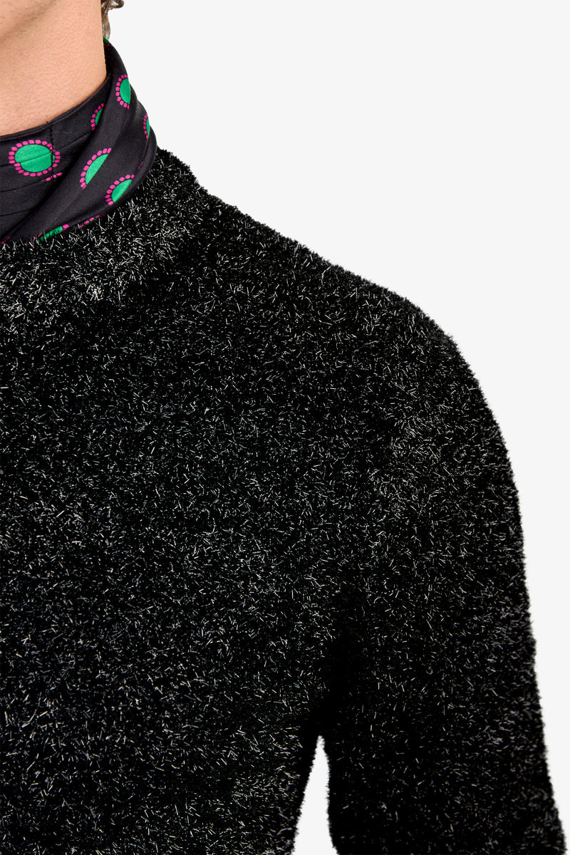 Close-up of a person wearing the ASTRON SPARKLE CREWNECK—a fuzzy black sweater—paired with a black scarf featuring green and pink circular patterns, against a plain white background.