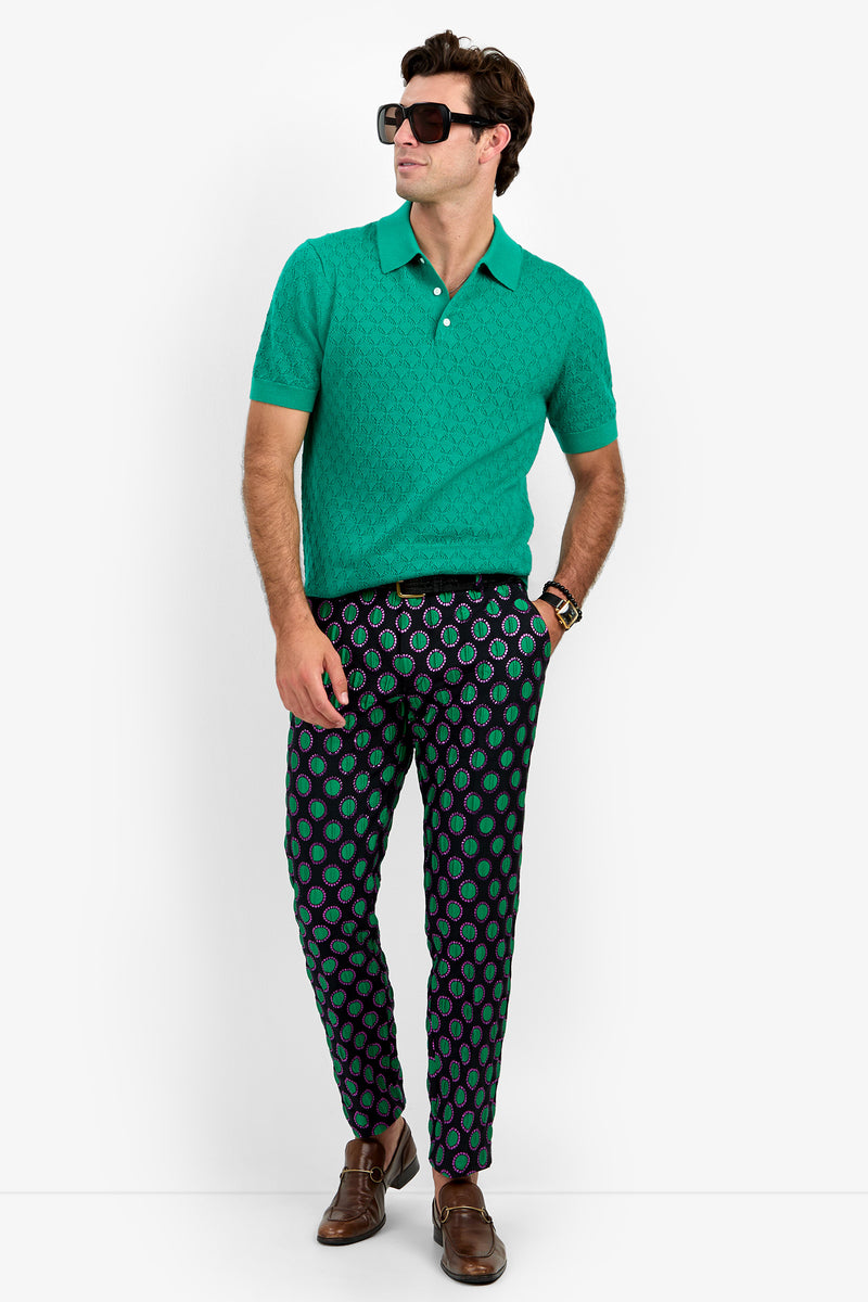 A man wearing dark sunglasses and the WAYLEN POLO—a green polo shirt—paired with patterned pants featuring green and purple shapes, stands with one hand in his pocket and looks to the side. He also wears brown dress shoes and a watch.