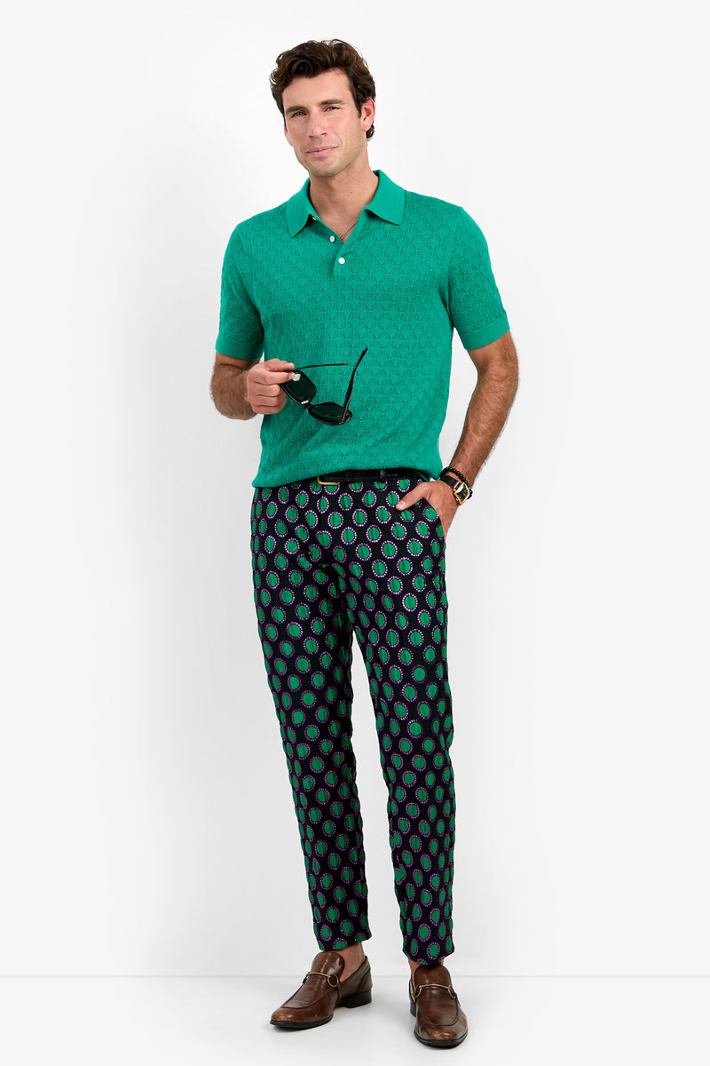 A man stands against a plain white background wearing the WAYLEN POLO, a green short-sleeve polo shirt, paired with patterned pants. He holds sunglasses in one hand and completes his look with brown dress shoes and a watch.