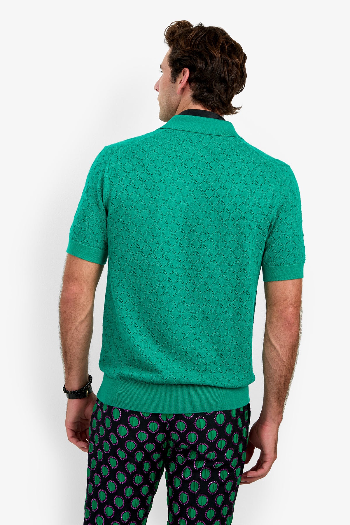 A man facing away, wearing the WAYLEN POLO—a textured green short-sleeve shirt—and patterned navy pants with green and magenta oval shapes, against a white background.