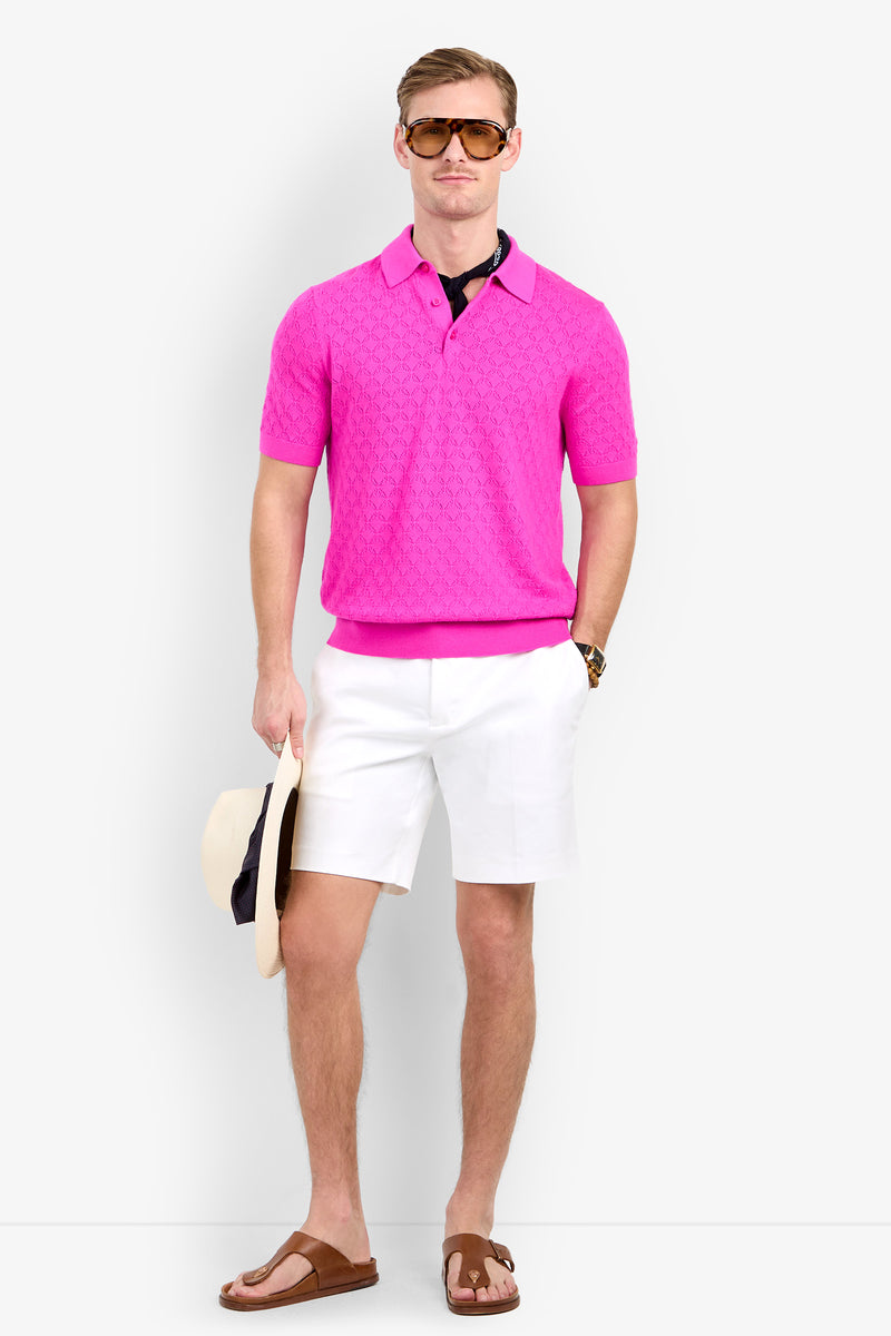 A man wearing a textured, short-sleeved, hot pink Waylen Polo, white shorts, brown sunglasses, and leather sandals, holding a straw hat at his side.