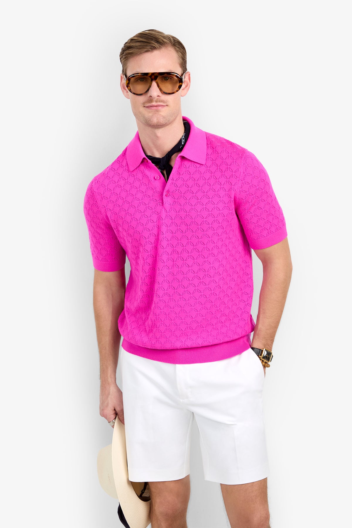 A man wears a bright pink Waylen Polo with a textured knit pattern, a black neckerchief, and white shorts.
