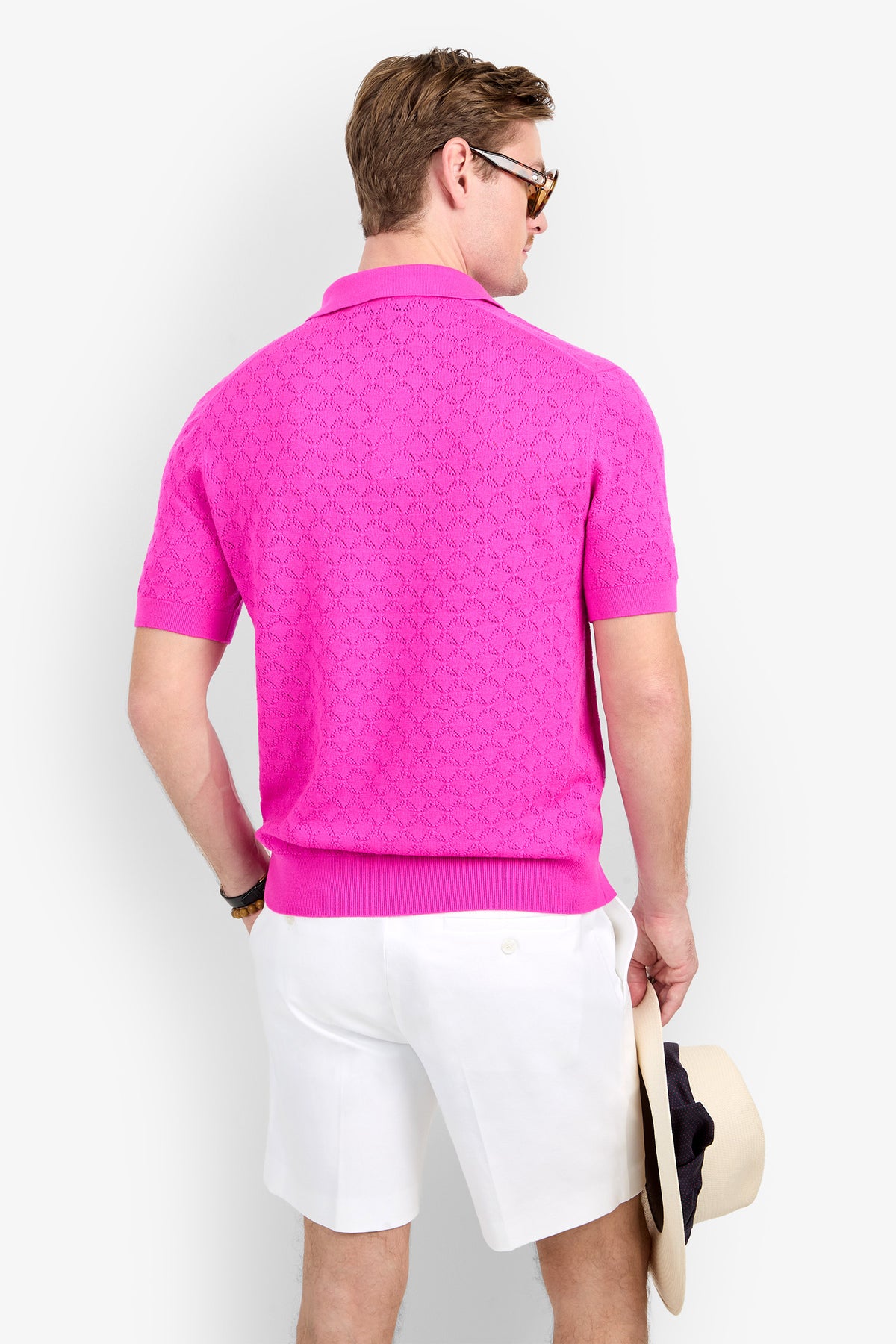 A man seen from behind wearing a bright pink, textured geometric Waylen Polo and white shorts, holding a straw hat in his hand.