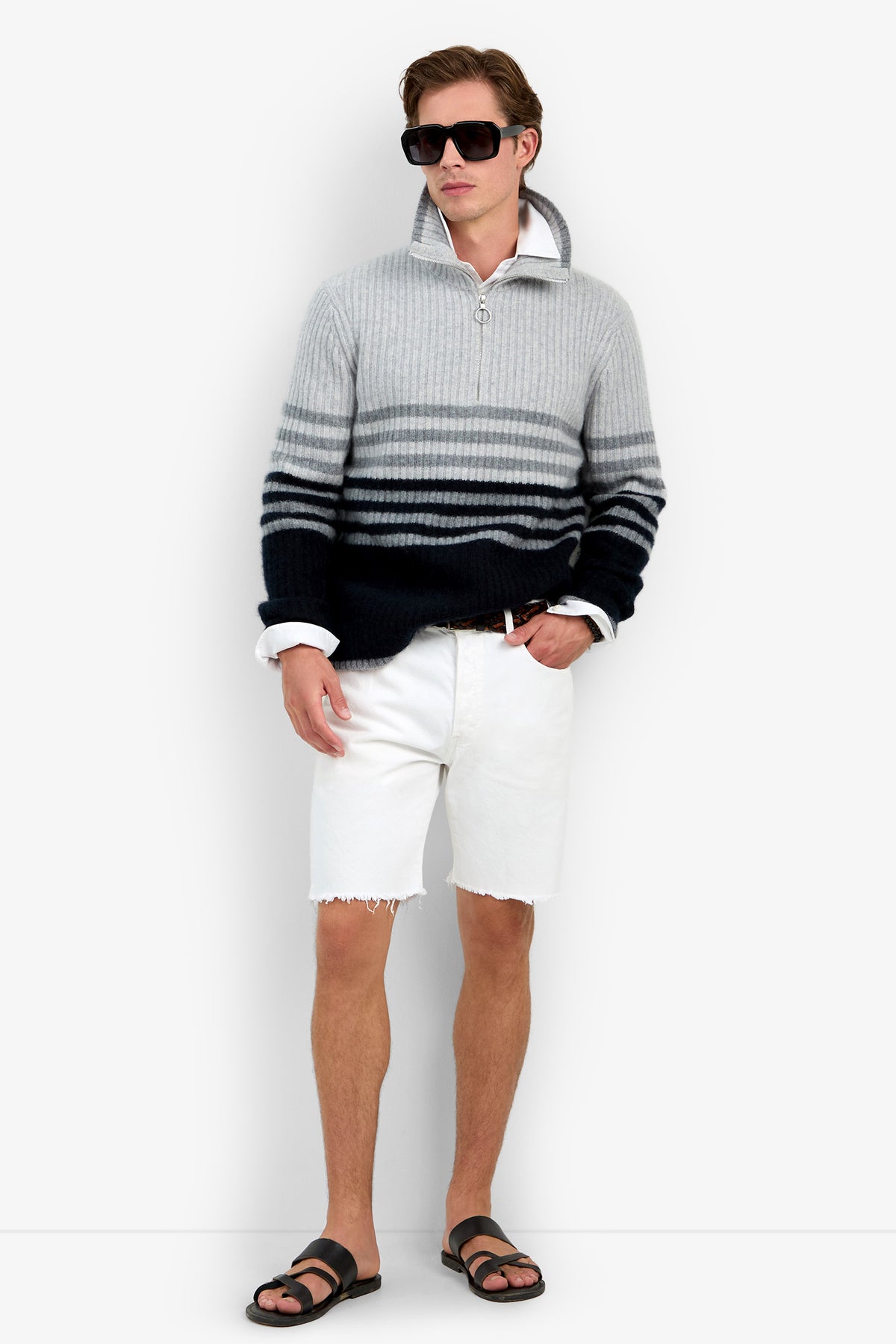 A man wearing the HEMMINGWAY QUARTER ZIP PULLOVER in GREY HEATHER, paired with a white collared shirt, white shorts, black sandals, and sunglasses stands against a plain background.